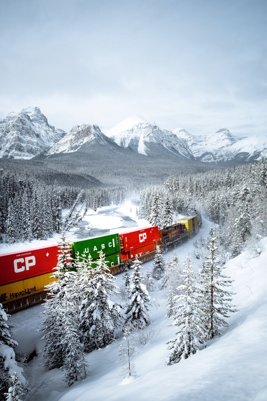 a moving train on a snow covered ground near the snow covered trees and mountains
