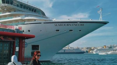 woman sitting by table on shore with cruise ship behind