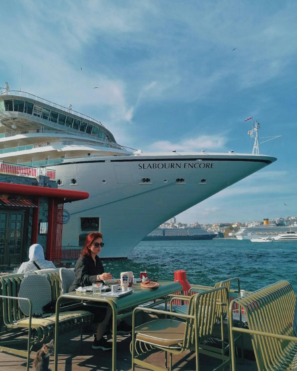 woman sitting by table on shore with cruise ship behind