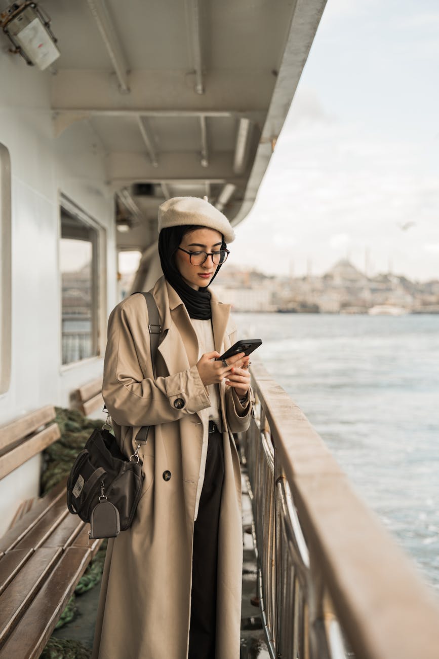 young woman in beige coat and white beret on black headscarf texting while traveling by ferry