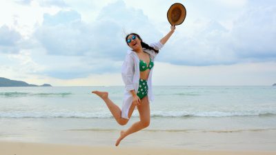 woman jumping on seashore and holding hat