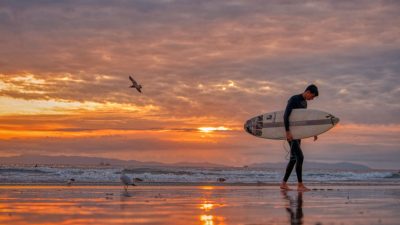 man walking on beach carrying white surfboard during sunset