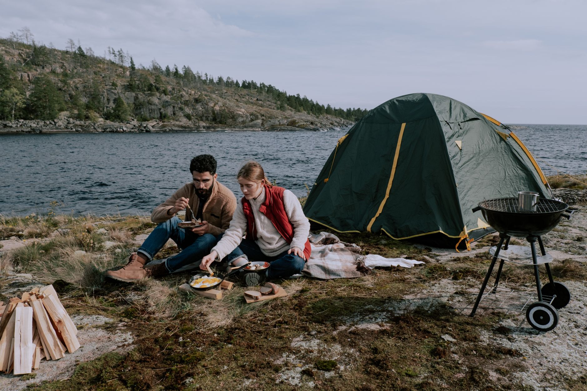 a couple sitting on picnic mat near a tent along a river