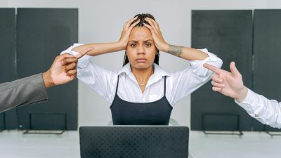 woman sitting in front of laptop with head in hands and men pointing at her