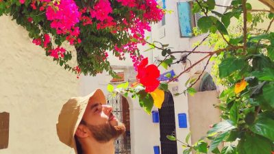man in black crew neck shirt with bucket hat smelling flowers in an alley