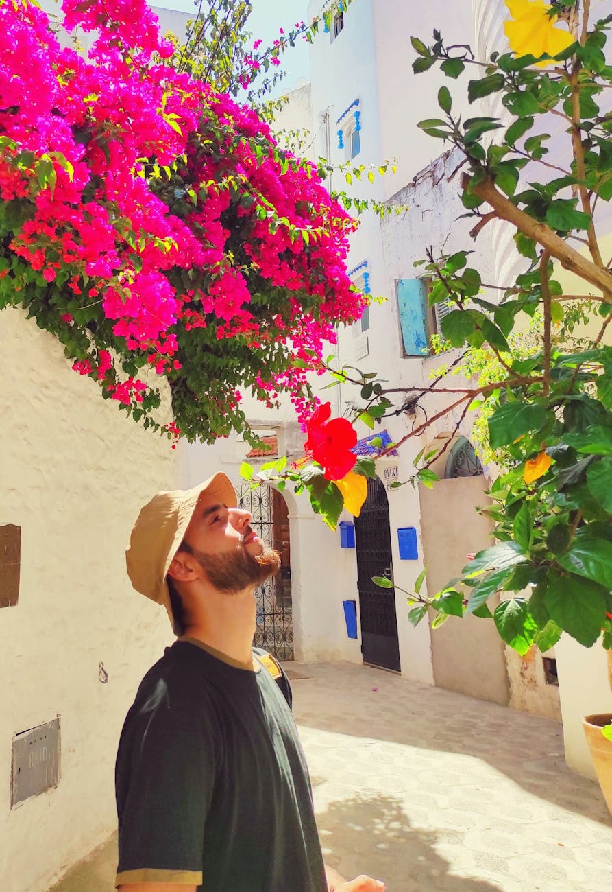 man in black crew neck shirt with bucket hat smelling flowers in an alley
