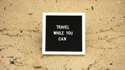 board with text travel while you can on the sand