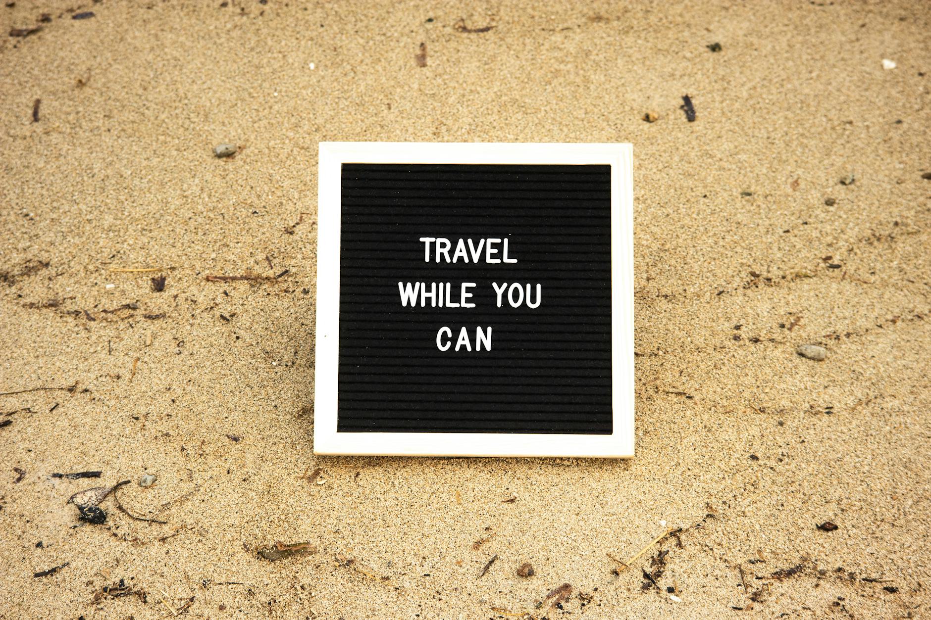board with text travel while you can on the sand